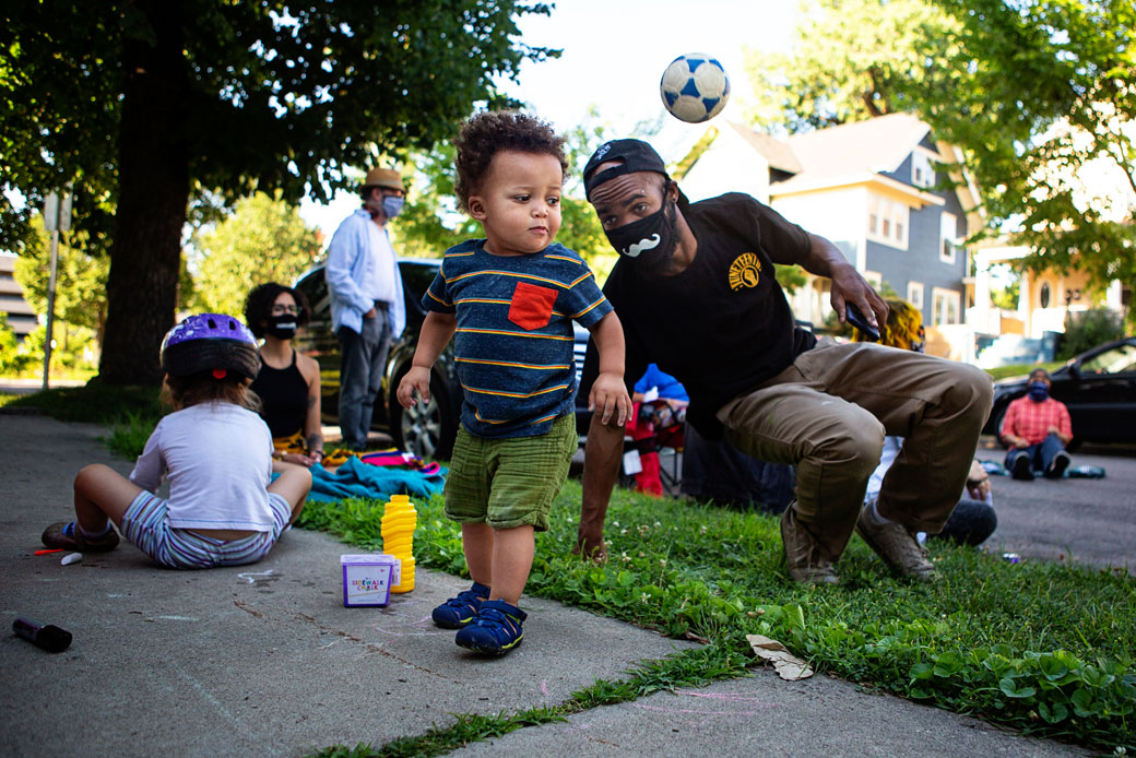 A toddler bounces a ball as his father keeps watch during a community conversation in Minneapolis, July 2020. (Getty/Los Angeles Times/Jason Armond)