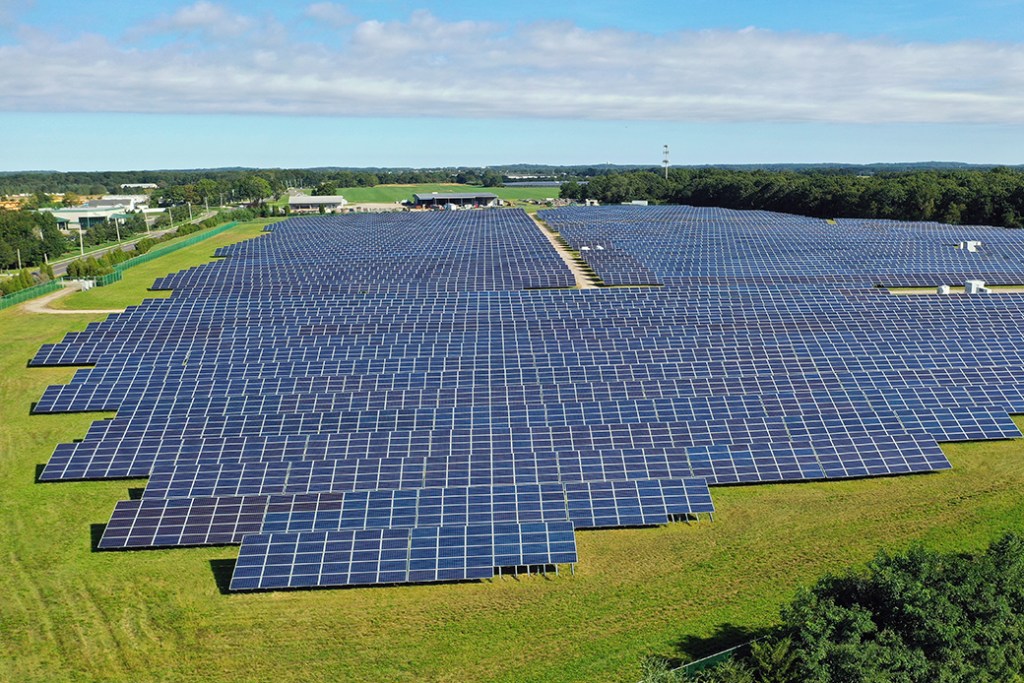 Aerial view of a green grassy field with a large array of blue solar panels, with trees off to the right of the frame.