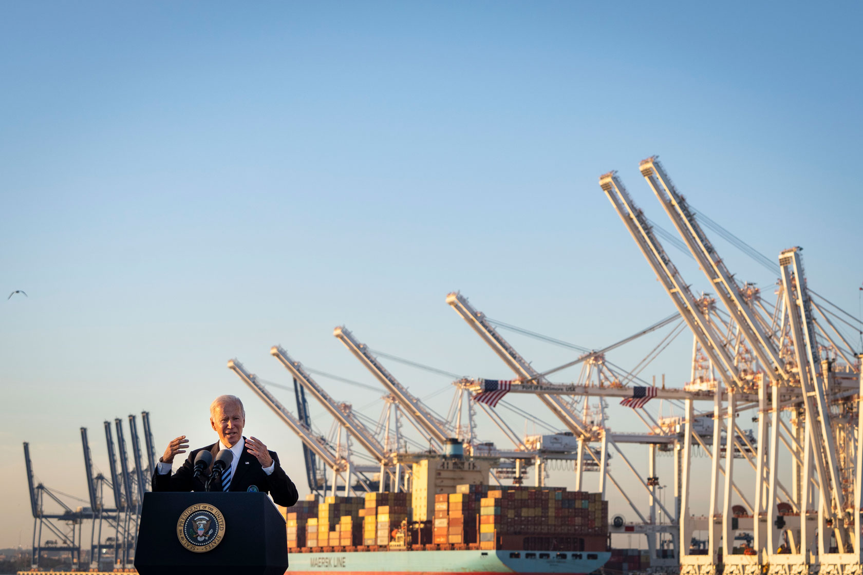 Photo shows President Joe Biden standing at a podium in front of a large port with a docked cargo ship.