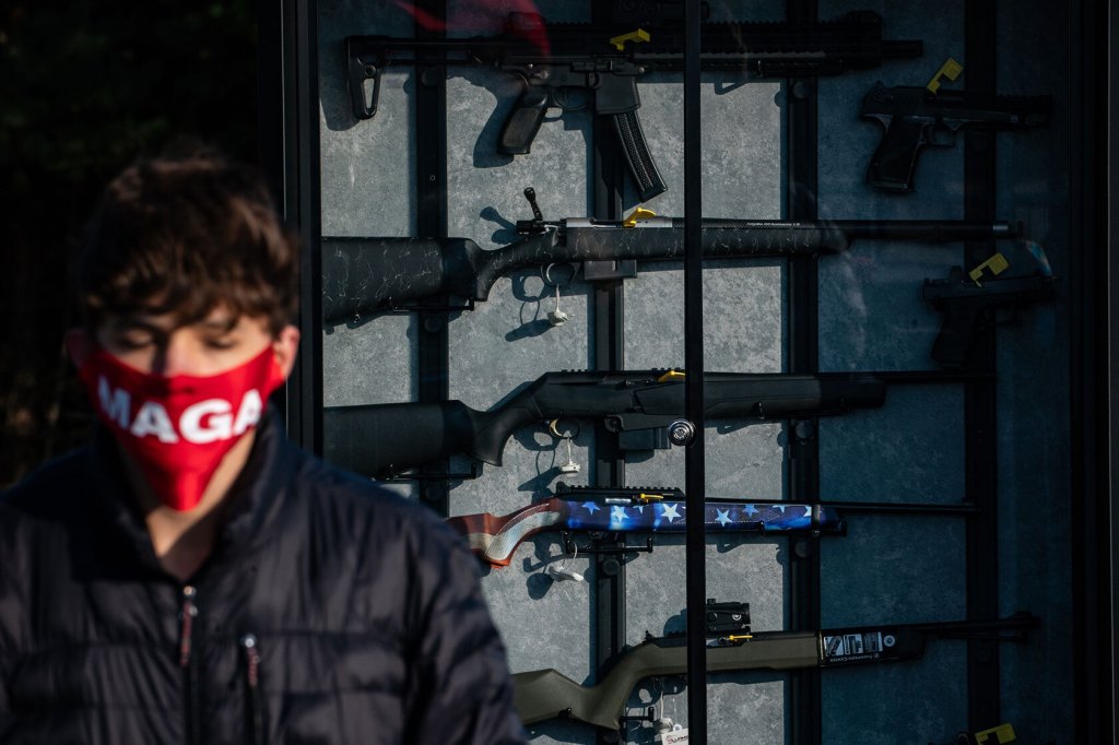 A rallier with a MAGA face mask stands in front of a gun display with semi-automatic rifles.