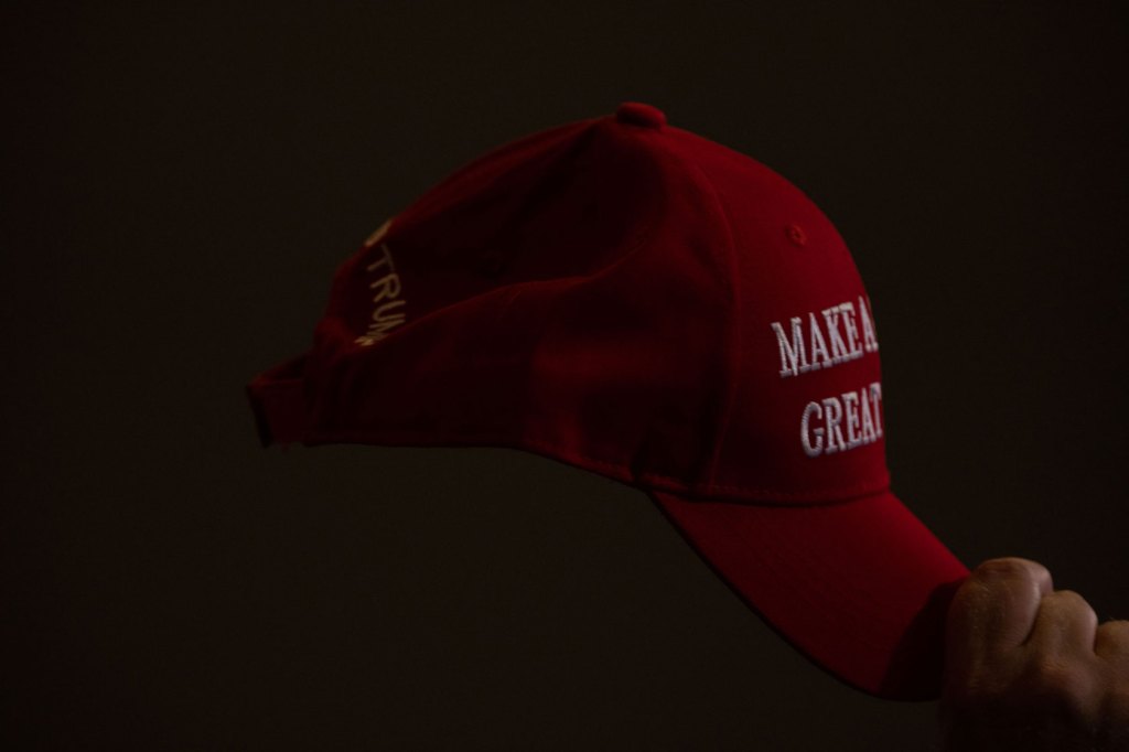A Trump supporter holds a MAGA hat with one hand on the brim.