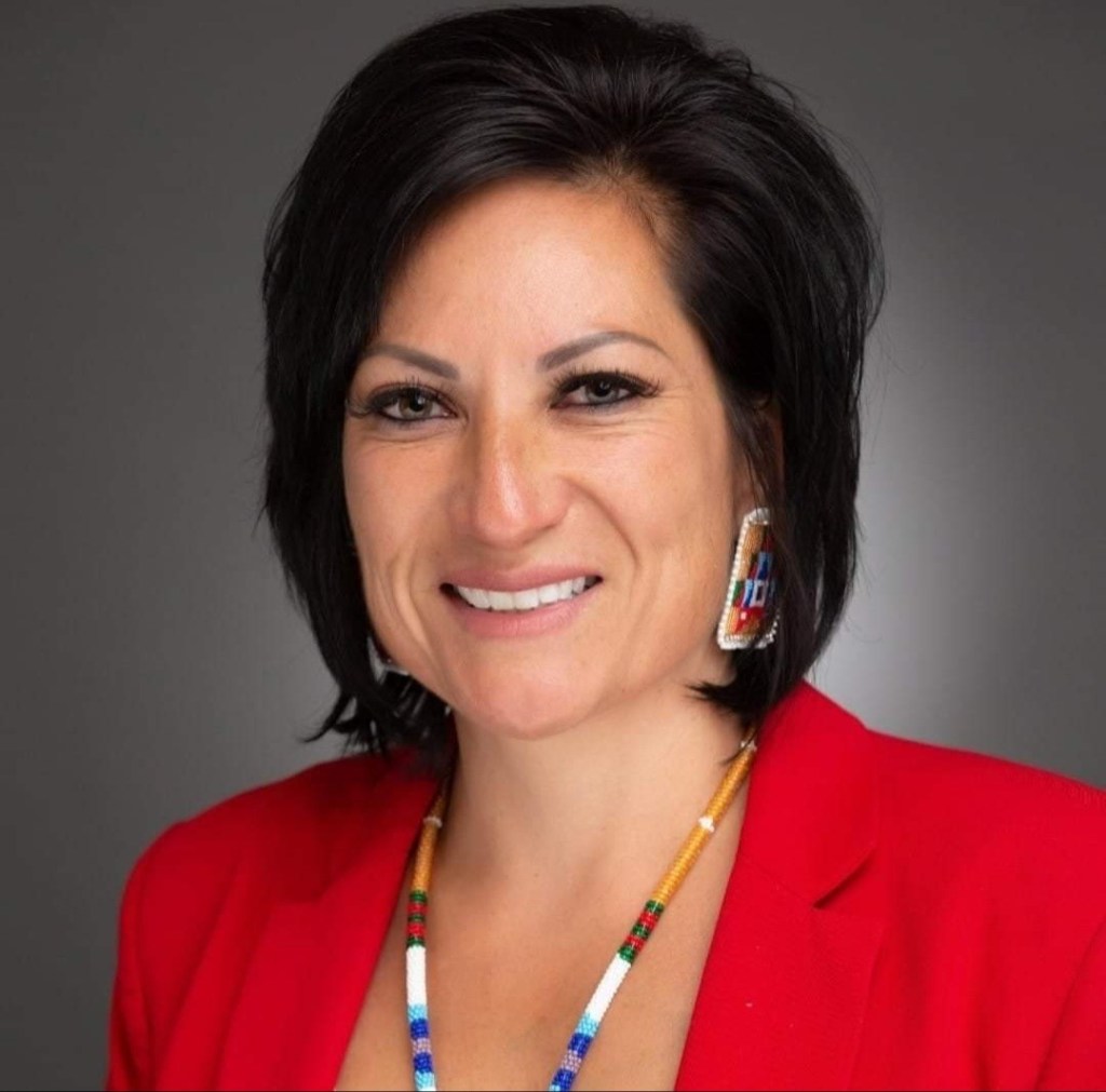 Photo shows a headshot of Amber Torres wearing a red blazer and colorful jewelry