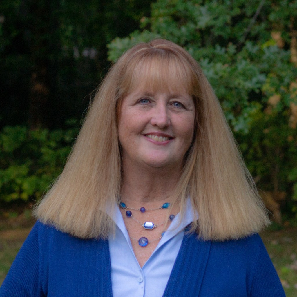 Photo shows Nancy Murphy smiling for a photo wearing all blue, with greenery in the background