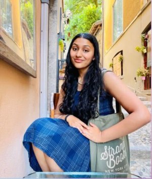 Photo shows Aarika Roy posing for a photo wearing a blue dress, with yellow buildings in the background
