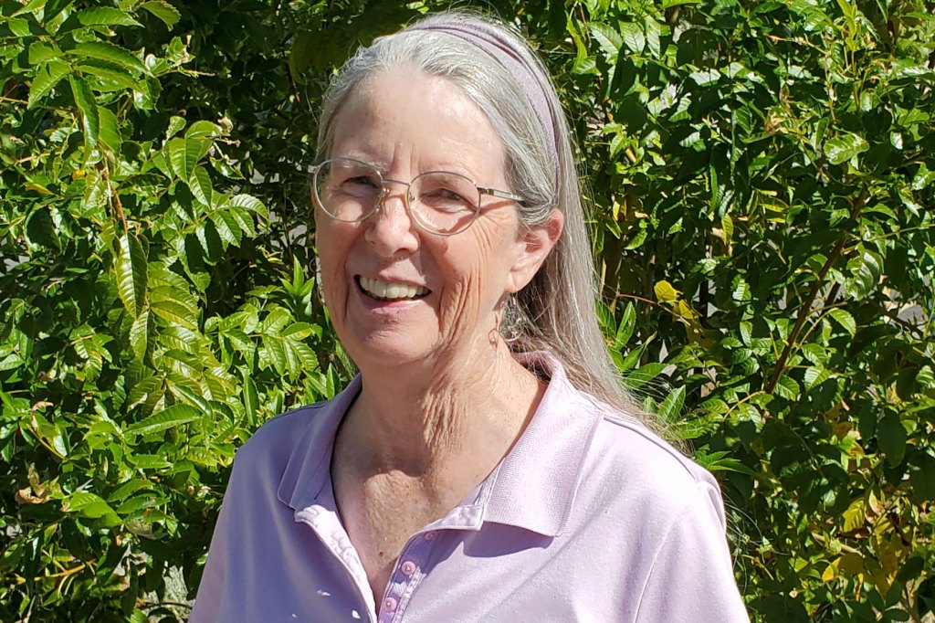 Photo shows Carol Brown smiling at the camera wearing a light purple shirt, with greenery in the background