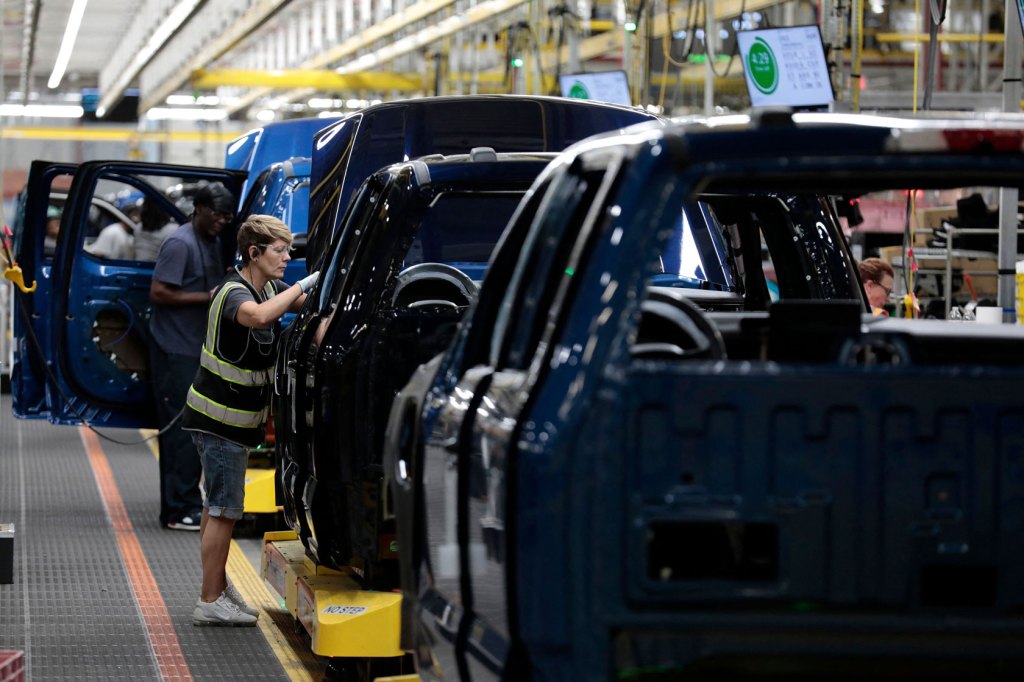 Photo shows a worker wearing a reflective vest working on a vehicle in an assembly line.