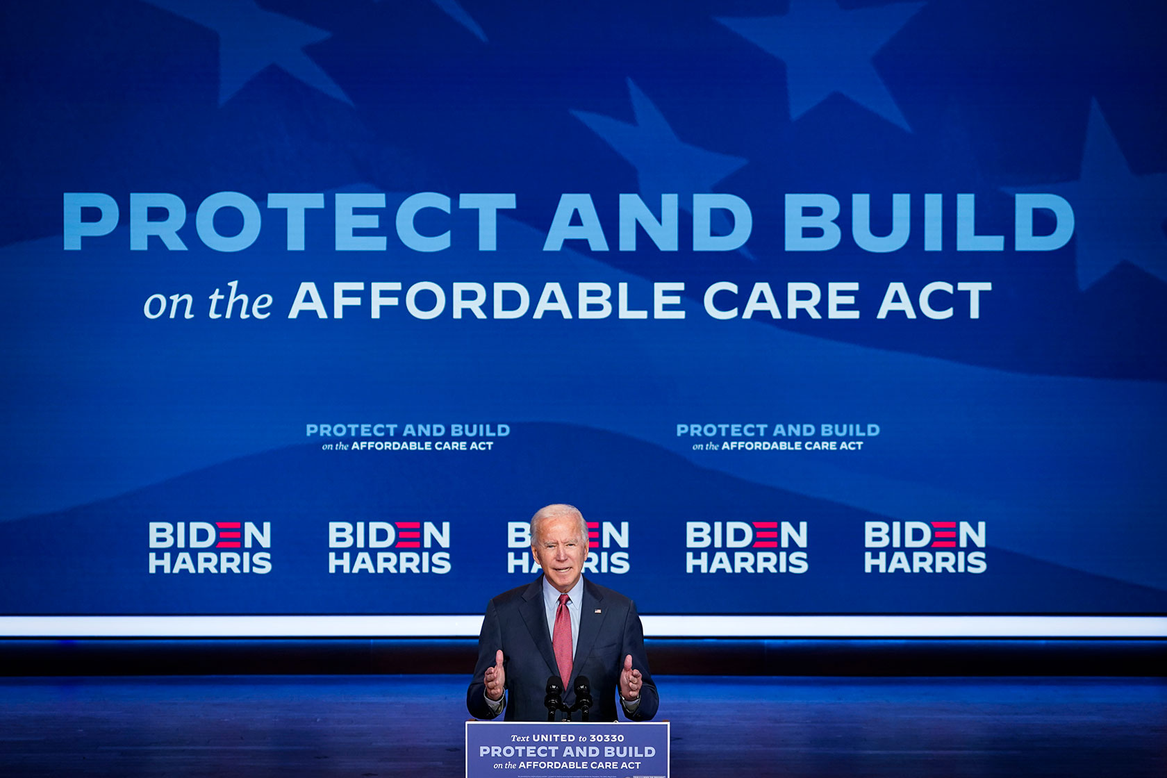 Photo shows Joe Viden giving a speech behind a podium, with a large blue sign in the background reading