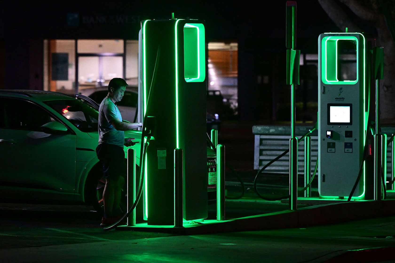 Photo shows a man plugging his car into a EV charger that glows green in the night