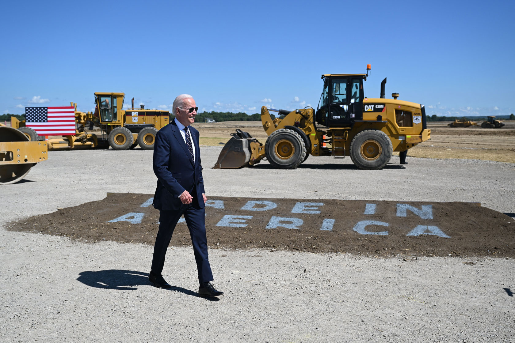 Photo shows Joe Biden walking in front of a construction site with yellow construction machinery in the background, and a 