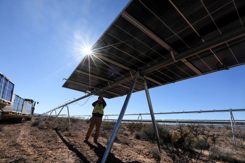 Photo shows a worker standing underneath a solar panel, reaching up to secure straps, in an open field on a sunny day