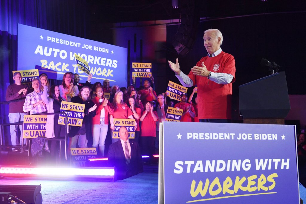 President Joe Biden is seen speaking from a podium in front of a crowd of people holding signs in support of the United Auto Workers union.