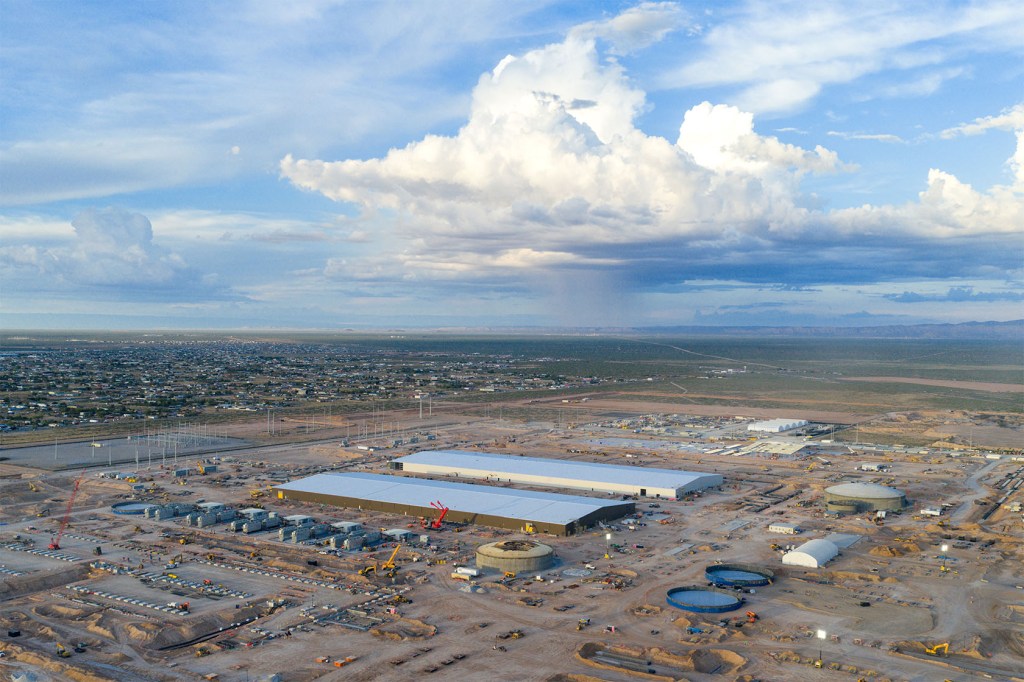 Aerial view of a data center construction site with two long warehouse buildings and storage tanks on cleared desert land, a town and large storm cloud in the distance.
