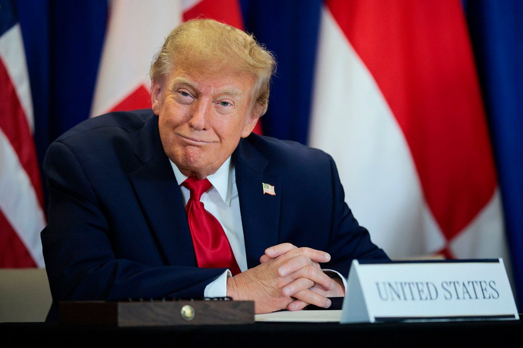 President Donald Trump, in a navy suit and red tie, sits at a table in front of a row of U.S. flags.